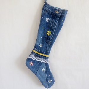 May include: A blue denim holiday stocking crafted from repurposed jeans, featuring a metal button at the top, a yellow decorative trim, white lace edging, and various floral patterns created with small colorful beads and pearls.