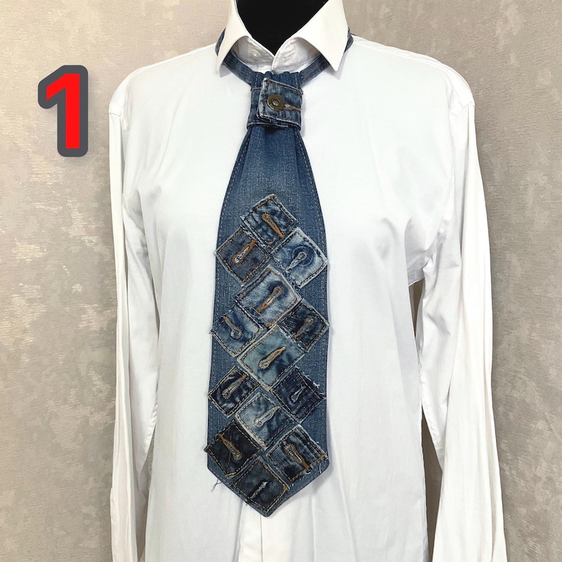 May include: A necktie constructed from blue denim fabric featuring a patchwork design of square denim pieces, each with a visible buttonhole. The tie is secured at the collar with a metal button and is worn over a crisp white long-sleeve button-down shirt.