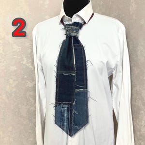 May include: A necktie constructed from various shades of blue denim fabric patches with visible frayed edges and exposed seams. The tie features a central metal button detail at the knot and is displayed against a crisp white collared shirt.