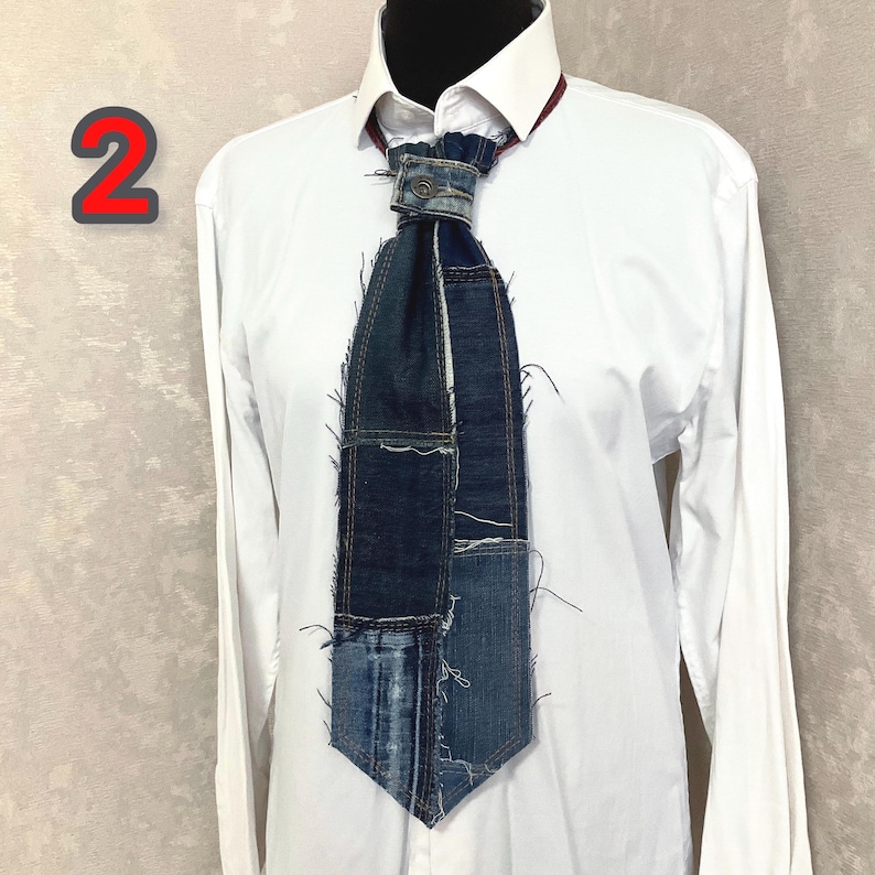 May include: A necktie constructed from various shades of blue denim fabric patches with visible frayed edges and exposed seams. The tie features a central metal button detail at the knot and is displayed against a crisp white collared shirt.