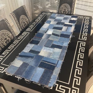 May include: A blue denim table runner made from patchwork squares of different shades of blue denim. The runner is on a black table with a white Greek key design border.