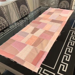 May include: A rectangular patchwork table runner in shades of pink, peach, and beige. The runner is made of various fabric squares sewn together. The table runner is placed on a black table with a decorative border.