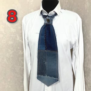 May include: A necktie crafted from various shades of blue denim fabric with frayed edges and visible stitching. The tie features a central metal button at the knot area and is displayed on a crisp white long-sleeved button-down shirt.