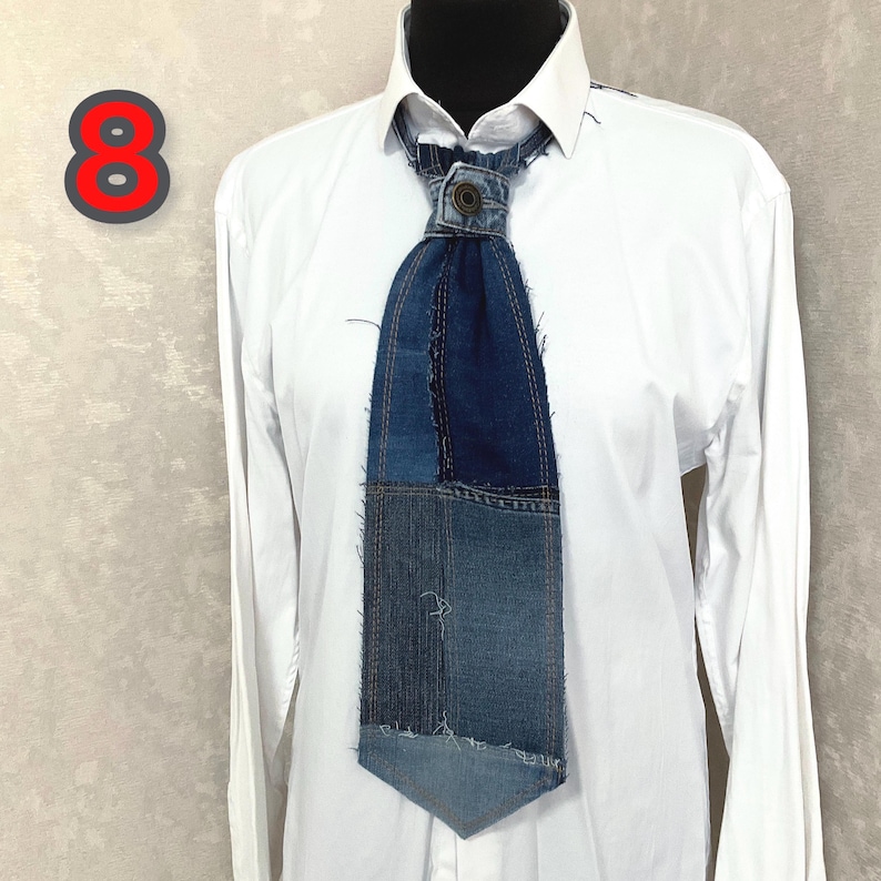 May include: A necktie crafted from various shades of blue denim fabric with frayed edges and visible stitching. The tie features a central metal button at the knot area and is displayed on a crisp white long-sleeved button-down shirt.