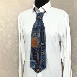 May include: A necktie constructed from repurposed blue denim fabric featuring multiple metal buttons, a vertical zip, and a tan leather patch with the text Levi Strauss & Co. San Francisco, Cal. Original Riveted Quality Clothing, worn over a white shirt.