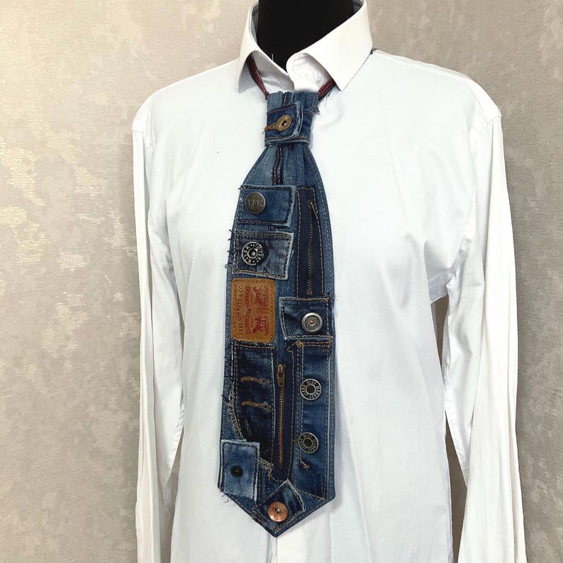 May include: A necktie constructed from repurposed blue denim fabric featuring multiple metal buttons, a vertical zipper, and a tan leather patch with the text Levi Strauss & Co. San Francisco, Cal. Original Riveted Quality Clothing, worn over a white shirt.