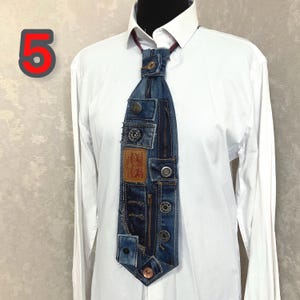 May include: A necktie crafted from repurposed blue denim fabric, featuring a prominent tan leather brand patch, multiple metal buttons, and a vertical zipper detail. The tie is displayed against a crisp white long-sleeved collared shirt.