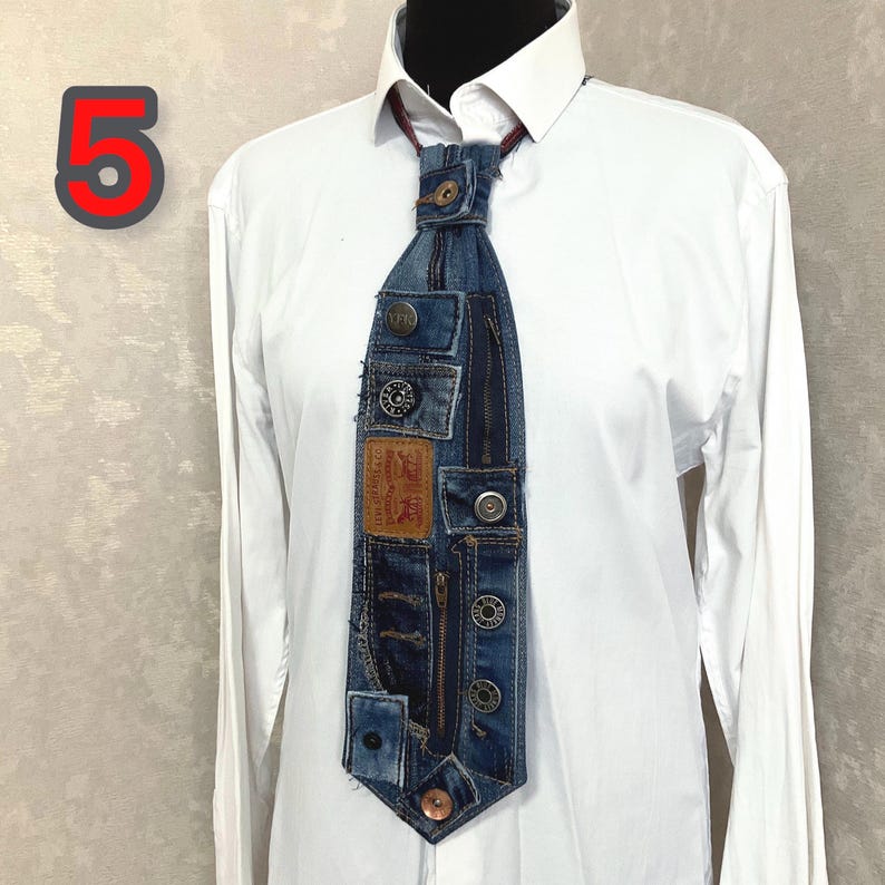 May include: A necktie crafted from repurposed blue denim fabric, featuring a prominent tan leather brand patch, multiple metal buttons, and a vertical zipper detail. The tie is displayed against a crisp white long-sleeved collared shirt.