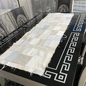 May include: A rectangular, patchwork table runner in shades of white, cream, and light blue. The runner is made of individual fabric squares stitched together. It is placed on a black table with a white Greek key design.
