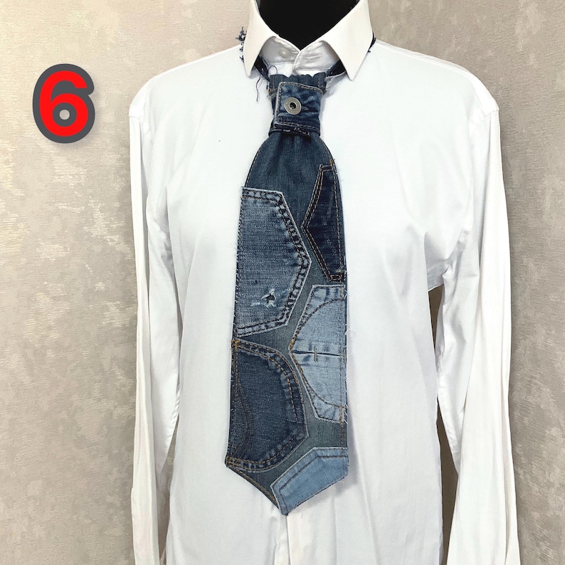 May include: A necktie constructed from various shades of blue denim fabric patches, featuring visible stitching, frayed edges, and a metal button at the knot. The tie is displayed against a plain white collared shirt with a red number 6 in the corner.