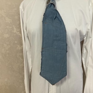 May include: A necktie crafted from light blue denim fabric featuring visible stitching lines and frayed edges. The tie is positioned against a plain white long-sleeved button-down shirt, showcasing a structured, multi-paneled design.