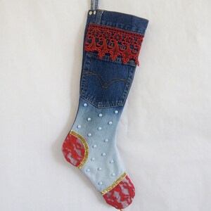 May include: A denim Christmas stocking with a blue and red color scheme. The stocking is made from repurposed jeans and features a red lace cuff and accents. The toe and heel are decorated with red lace and gold trim, and it is embellished with small white pearls.