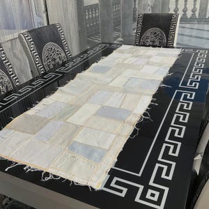 May include: A rectangular patchwork table runner in shades of white and light blue. The runner is made of various fabric squares sewn together. It rests on a black table with a white geometric border. The table is surrounded by chairs with a black and white design.