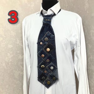 May include: A necktie constructed from dark blue denim fabric patches, featuring a central knot with a metal button and a series of various metallic buttons arranged in a grid pattern down the length of the tie, worn over a crisp white shirt.