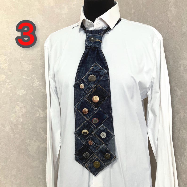 May include: A necktie constructed from dark blue denim fabric patches, featuring a central knot with a metal button and a series of various metallic buttons arranged in a grid pattern down the length of the tie, worn over a crisp white shirt.
