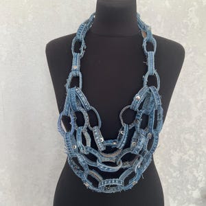 May include: A unique denim necklace with a layered design. The necklace is constructed from strips of blue denim, connected by metal rings and rivets. The necklace is displayed on a black mannequin.