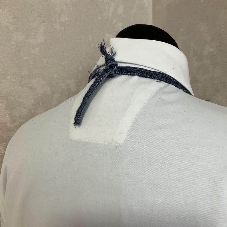 May include: A white collared shirt featuring a dark blue denim-like cord or strap tied in a knot at the back of the neck. The shirt has a structured collar and a reinforced fabric panel at the upper back, showcasing a clean and simple design.