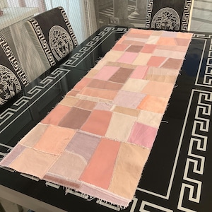 May include: A rectangular patchwork table runner in shades of pink, peach, and beige. The runner is made of fabric squares sewn together. The table it rests on is black with a white geometric border. The chairs have a Medusa head design.