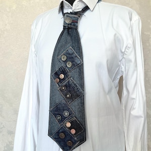 May include: A necktie crafted from dark blue denim fabric, featuring multiple square denim patches layered across the front. The tie is adorned with several metallic buttons and rivets, and it is worn over a crisp, long-sleeved white button-down shirt.