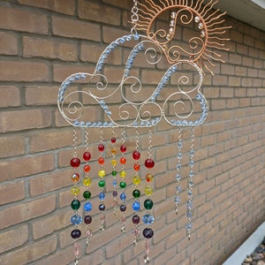 May include: A wire sculpture of a cloud with a sun above it. The cloud is decorated with blue beads and the sun is decorated with clear beads. The sculpture has rainbow coloured beads hanging from it.