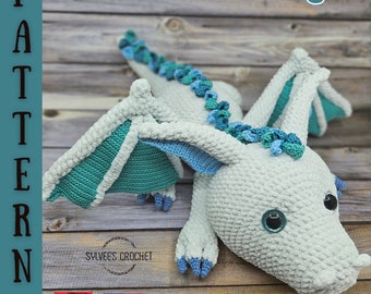 May include: A crocheted grey dragon with teal wings and a teal and blue tail. The dragon is lying on a wooden surface. The text "Stiria the dragon" is written above the dragon. The text "PATTERN PDF" is written on the left side of the image. The text "Sylvee's Crochet" is written below the dragon. The flags of Hungary and the United States are shown below the text.