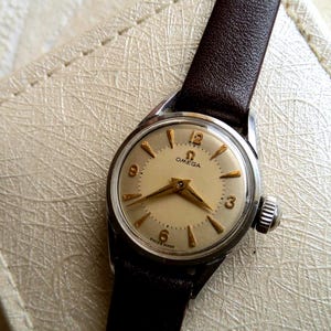 Könnte beinhalten: Eine Vintage-Armbanduhr von Omega mit weißem Zifferblatt, goldfarbenen Zeigern und Stundenmarkierungen sowie einem braunen Lederarmband.