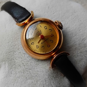 May include: A gold-tone wristwatch with a black leather strap. The watch face is round and has a gold-tone bezel. The watch face has black Roman numeral hour markers and a red second hand. The watch face also has the text "16 jewels" printed on it.