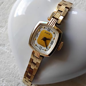 May include: A gold-toned wristwatch with a square face and a white dial. The watch has Roman numeral hour markers and a gold-coloured second hand. The watch face also has the text "ЗАРЯ" and "17 камней".