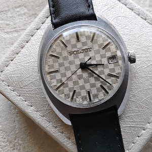 Könnte beinhalten: Eine Vintage-Armbanduhr von Poljot mit einem silberfarbenen Zifferblatt im Schachbrettmuster, schwarzen Stunden- und Minutenzeigern, einem dünnen Sekundenzeiger und einem Datumsfenster auf der Drei-Uhr-Position. Die Uhr hat ein silbernes Gehäuse und ein schwarzes Lederarmband.