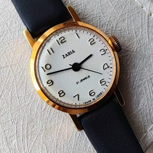 May include: A gold-tone wristwatch with a white face and black leather strap. The watch face has black hands and black Roman numeral hour markers. The watch face also has the text "Zaria" and "21 Jewels" printed on it.
