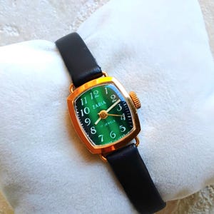 May include: A gold-tone wristwatch with a green face and black leather strap. The face has gold-tone hands and markers, and the text "Zaria 17 Jewels" is printed on the face.