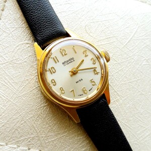 Puede incluir: Un reloj de pulsera Atlantic Miss vintage dorado con esfera crema y correa de cuero negro. La esfera del reloj presenta números y agujas doradas, con el nombre de la marca y "17 Jewels" impresos.