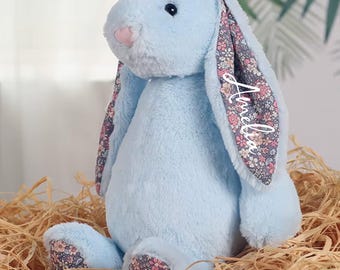 Op de afbeelding: Een lichtblauw pluche konijn met bloemmotief oren en voetkussentjes. Het konijn heeft een roze neus en de naam "Amelia" staat op één oor. Het konijn zit op een bedje van stro.