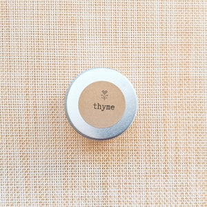 May include: A small, round, silver tin with a tan label. The label features a heart and floral design above the word "thyme" in a simple, black font. The tin is centered on a woven, tan surface.