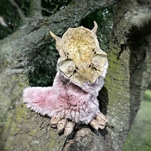 May include: A pink and brown plush creature with a skeletal head and long, clawed fingers. The creature is sitting on a tree branch.