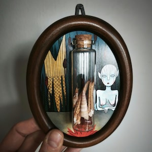 May include: A framed artwork featuring a glass bottle with a cork stopper, filled with beige objects, set against a painted backdrop. The artwork is enclosed in a dark brown oval frame, with a stylized figure on the right.