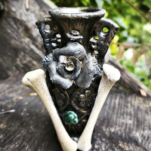 May include: A dark grey decorative vase with a rose design and two bone-shaped accents. The vase has a textured surface and a small green gem. The background is blurred with green foliage.