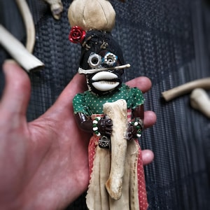 Puede incluir: Muñeca artesanal con un gorro negro, boca de concha y ojos de hardware reutilizado. La muñeca lleva una blusa verde con lunares y una falda beige con lunares rojos. Sostiene un hueso y tiene una pequeña rosa roja y un paquete de tela en la cabeza.