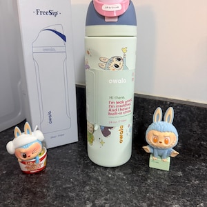 Puede incluir: Una botella de agua Owala FreeSip verde claro con tapa azul y rosa, con gr&aacute;ficos de conejos de dibujos animados y el texto "Hi there. I'm insulated. And I have a built-in straw." La botella es de 710 ml. Tambi&eacute;n se muestra una caja y dos figuras de conejos.