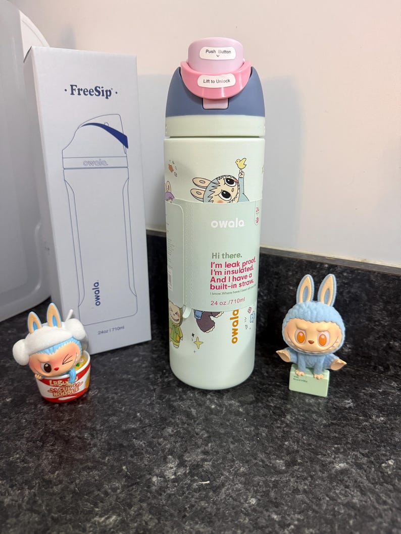 Puede incluir: Una botella de agua Owala FreeSip verde claro con tapa azul y rosa, con gr&aacute;ficos de conejos de dibujos animados y el texto "Hi there. I'm insulated. And I have a built-in straw." La botella es de 710 ml. Tambi&eacute;n se muestra una caja y dos figuras de conejos.