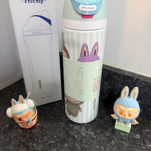 Puede incluir: Una botella de agua Owala FreeSip verde menta y blanca con gr&aacute;ficos de conejos, un bot&oacute;n azul y una caja a juego. Tambi&eacute;n hay dos peque&ntilde;as figuras de conejos.