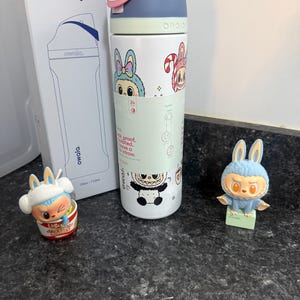Puede incluir: Una botella de agua Owala FreeSip blanca con gr&aacute;ficos de conejos de dibujos animados, una tapa rosa y azul y una caja a juego. Tambi&eacute;n hay dos figuras de conejos presentes, una en un cuenco y otra de pie.