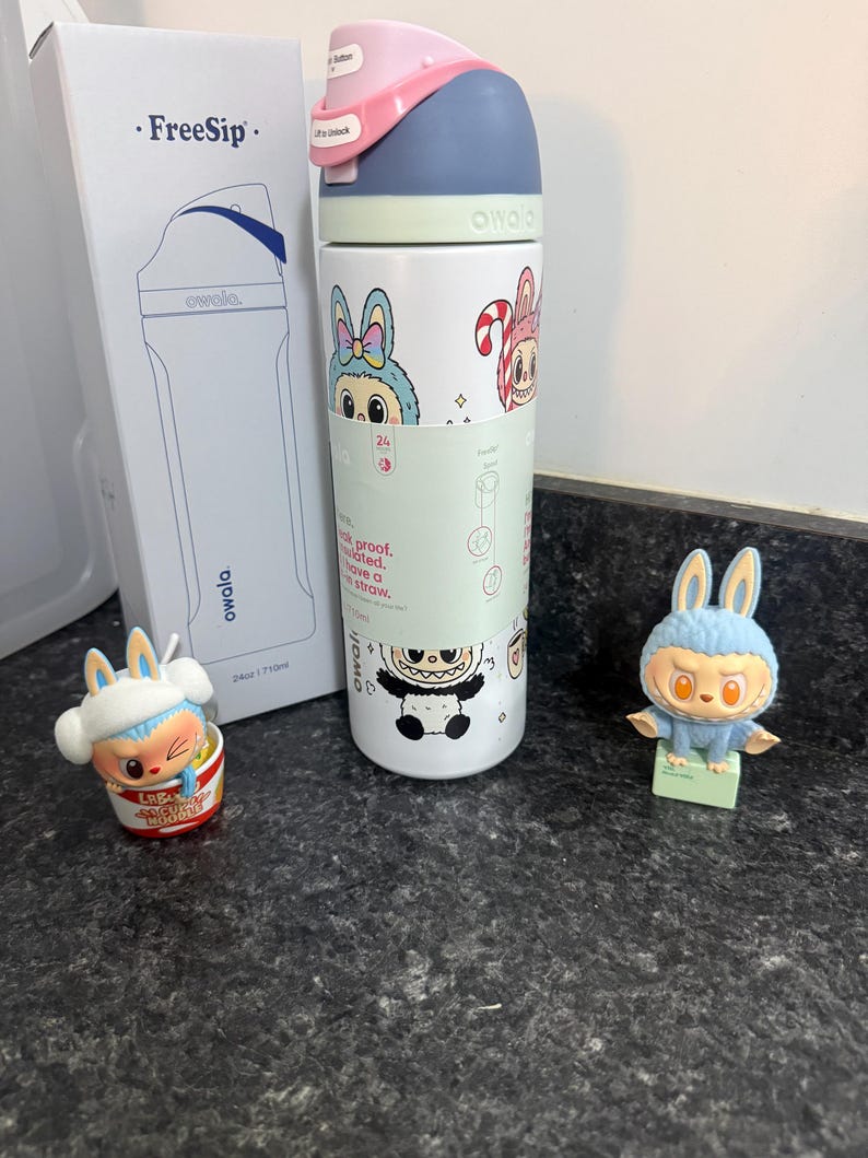 Puede incluir: Una botella de agua Owala FreeSip blanca con gr&aacute;ficos de conejos de dibujos animados, una tapa rosa y azul y una caja a juego. Tambi&eacute;n hay dos figuras de conejos presentes, una en un cuenco y otra de pie.