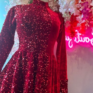 Puede incluir: Vestido rojo con lentejuelas, mangas largas y cuello alto. El corpiño y las mangas están cubiertos de lentejuelas, mientras que la falda es de material aterciopelado. El vestido se exhibe en un maniquí.