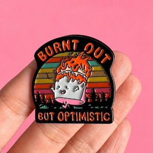 Peut inclure: Une broche en émail noir représentant une guimauve dessin animé avec une flamme sur la tête, sur un fond de coucher de soleil rétro avec des silhouettes d'arbres. Le texte BURNT OUT figure en haut et BUT OPTIMISTIC en bas en orange.