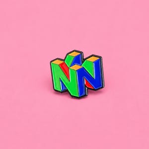 May include: A metal enamel pin shaped like a stylised letter N, featuring a three-dimensional block design with vibrant sections of bright green, royal blue, orange, and red, all outlined in a bold black border against a solid pink background.