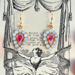 May include: Gold-tone earrings with a teardrop-shaped, pink-red center stone surrounded by light blue crystals. The earrings hang from a gold hook. The card behind the earrings has a vintage illustration and text that reads "REGENCY GLAM".