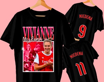 May include: Black t-shirts featuring a portrait of Viviane Miedema, with the name in pink and white. One shirt has the name and number 9 in red, and another has the number 11. The shirts are hanging on wooden hangers.