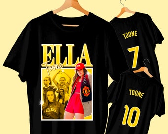 May include: Black t-shirt with a gold graphic of a woman soccer player with the text "Ella Toone" and a soccer ball. The shirt also features a group of women soccer players in the background.