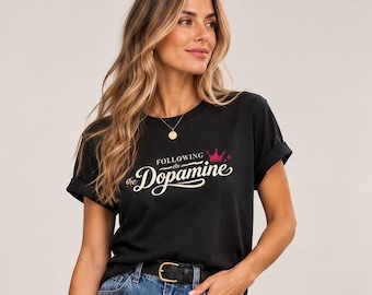 Könnte beinhalten: Ein schwarzes Kurzarm-T-Shirt mit dem Text "FOLLOWING the Dopamine" und einer kleinen rosa Kronengrafik, kombiniert mit einer blauen Jeans, einem schwarzen Gürtel, einer dunkelroten geflochtenen Handtasche und dunkelroten Pantoletten.